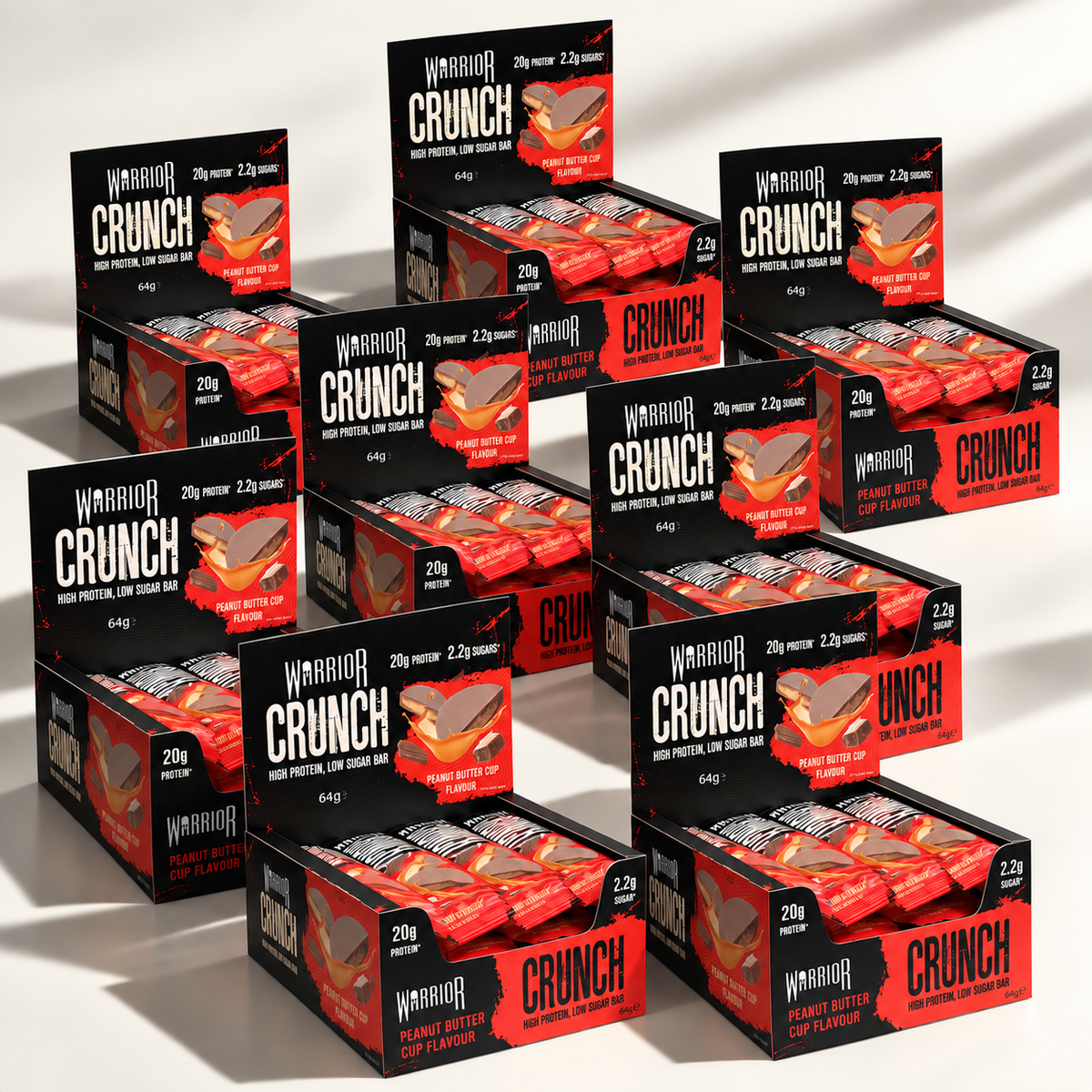 Eight Warrior CRUNCH Peanut Butter Cup protein bar display boxes arranged in an overlapping cluster on an off-white background, with red and black packaging and dramatic studio lighting.