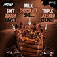 Warrior Crunch Fudge Brownie protein bar, cut open to show the soft dough layer, milk chocolate shell and triple-layered crunch with a caramel centre, wrapper partially peeled back on a dark brown background with chocolate crumb pieces.