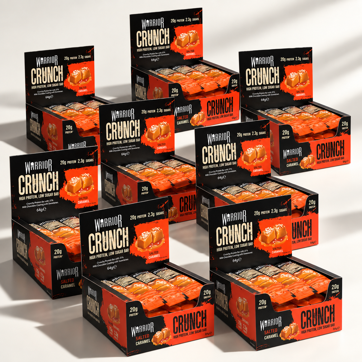 Eight Warrior CRUNCH Salted Caramel protein bar display boxes arranged in an overlapping cluster on an off-white background, with red and black packaging and dramatic studio lighting.